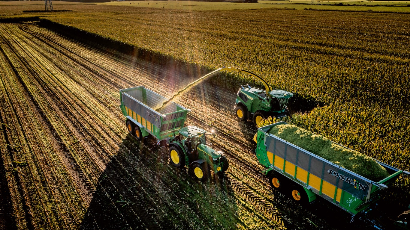 A John Deere self-propelled forage harvester operating in a cornfield with corn coming out of the spout and filling a wagon being towed by a john Deere tractor.