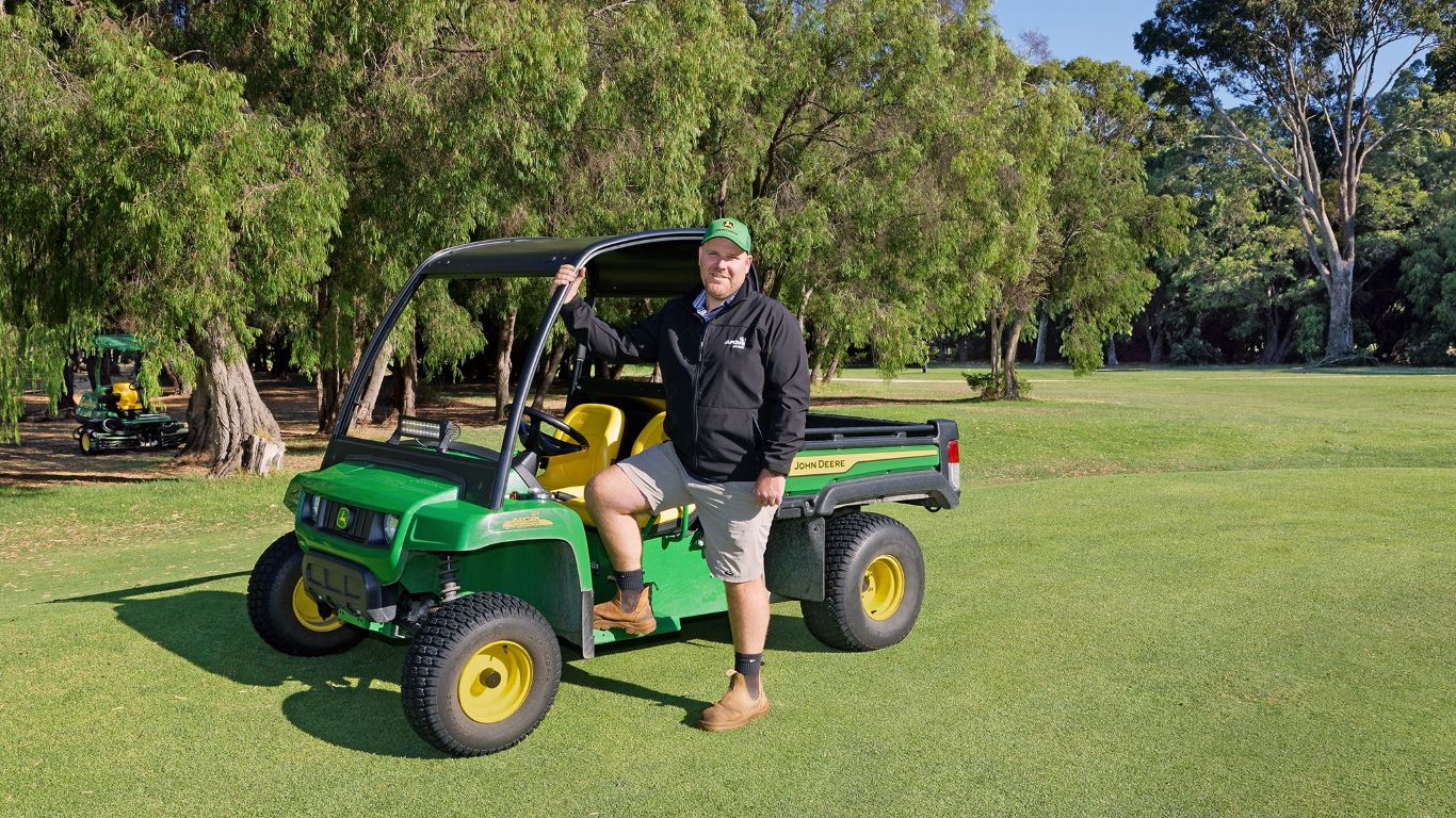 A person standing in front of a green utility vehicle on lawn with trees in the background.