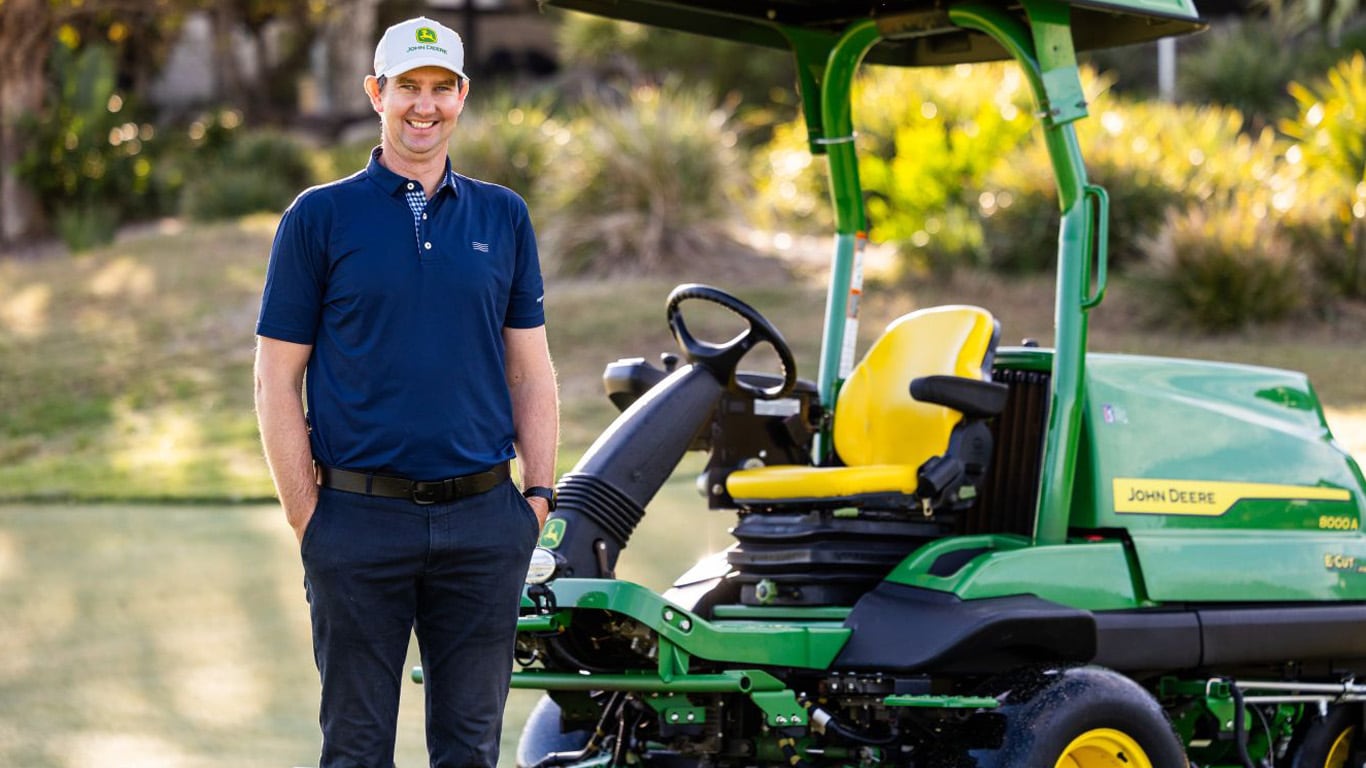 A man wearing a blue polo shirt, dark jeans a baseball cap in front of a green professional fairway mower.