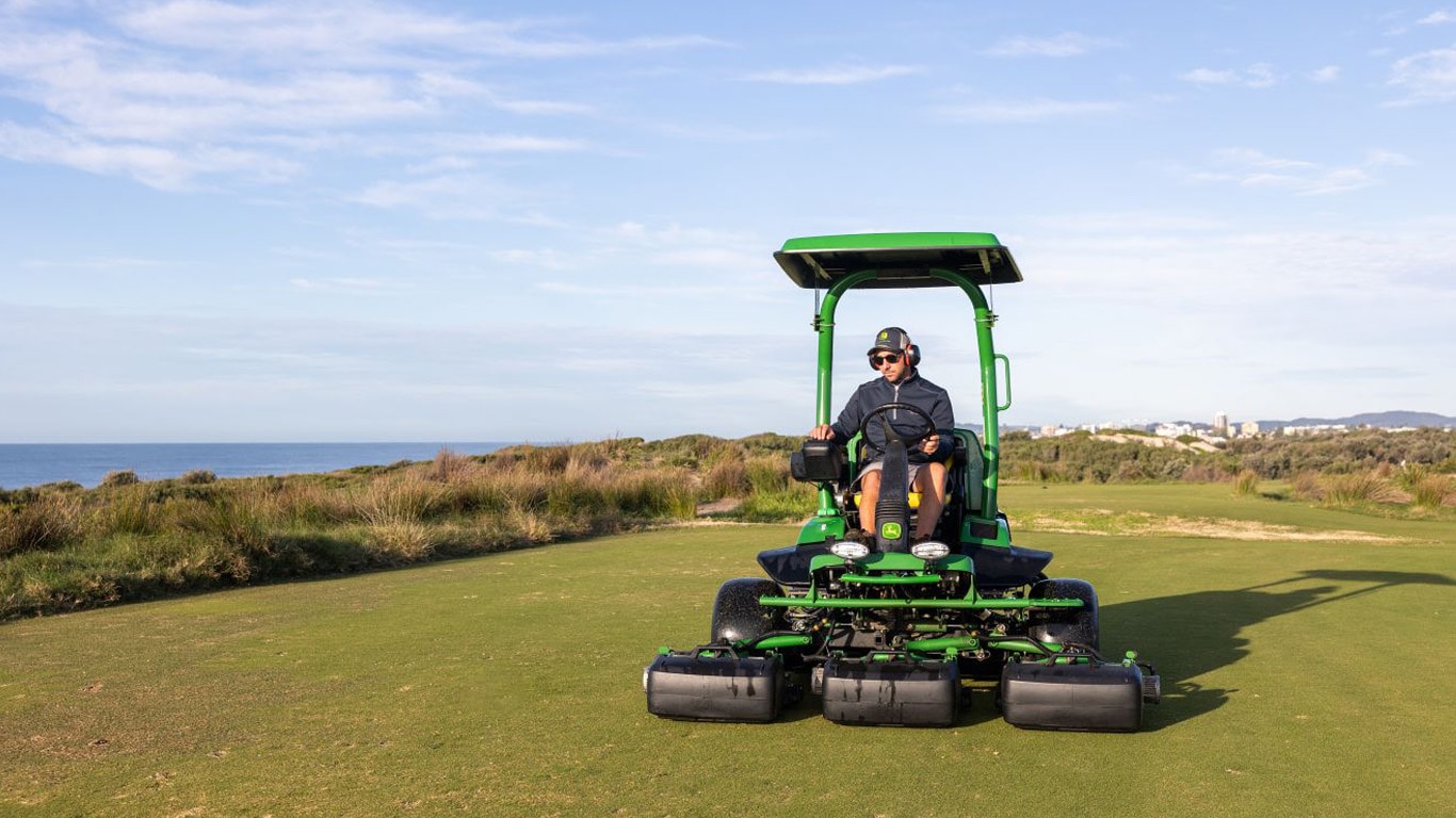 A man driving a green professional fairway mower on a golf course with the ocean in the background.