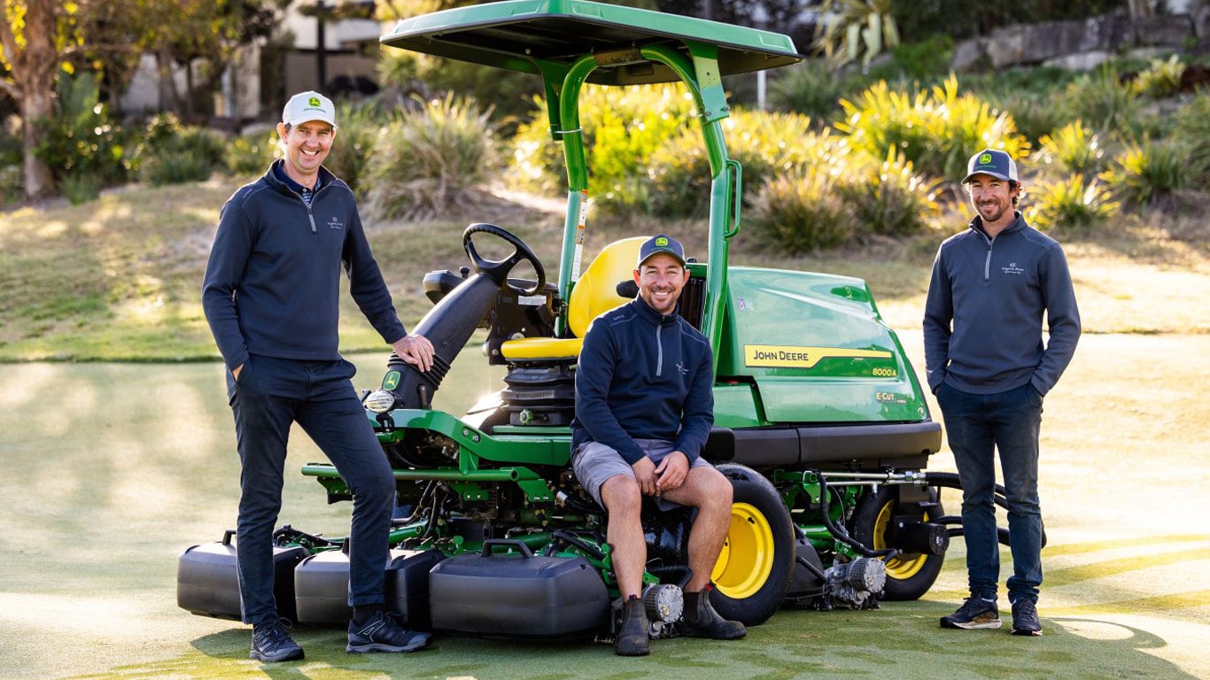 Three men wearing blue jumpers and baseball caps with a green fairway mower on a golf course.