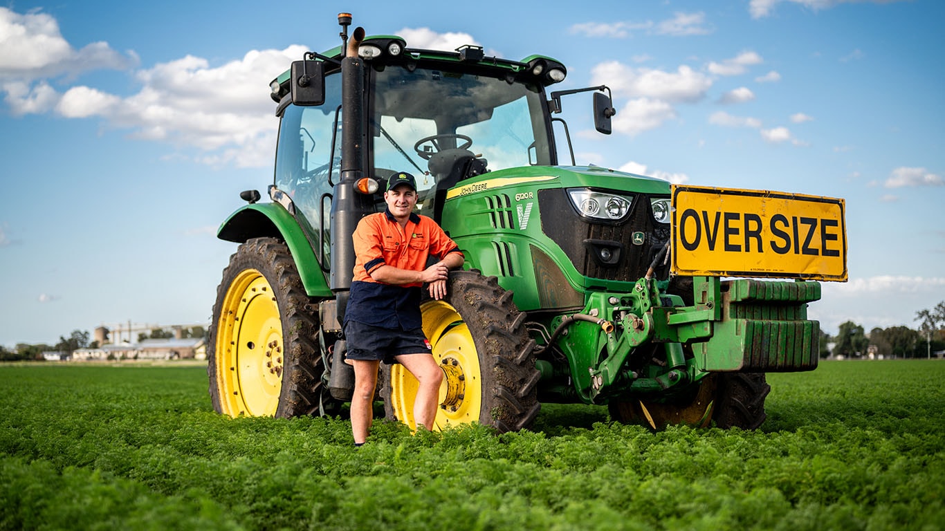 Mitch Brimblecombe standing in carrot farm, leaning on John&nbsp;Deere 6120R tractor