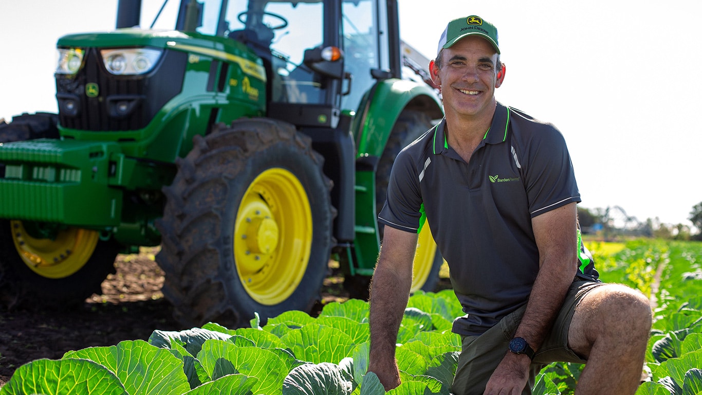A man wearing a cap kneels on one knee in a cabbage field, with a tractor parked in the background.