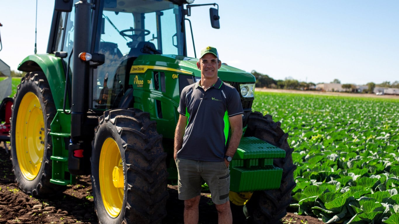 A man wearing a cap stands with his hands in his pockets in front of a parked tractor beside a cabbage field.