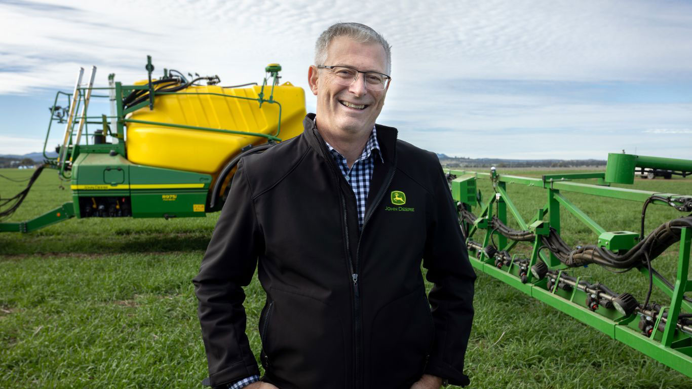 Man in black jacket in front of green and yellow machine in field with green ground and blue sky