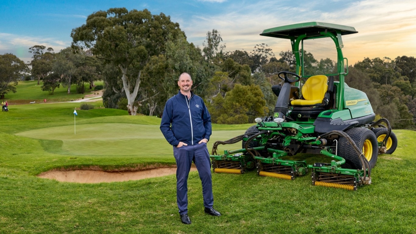 Man and John Deere mower beside golf green.