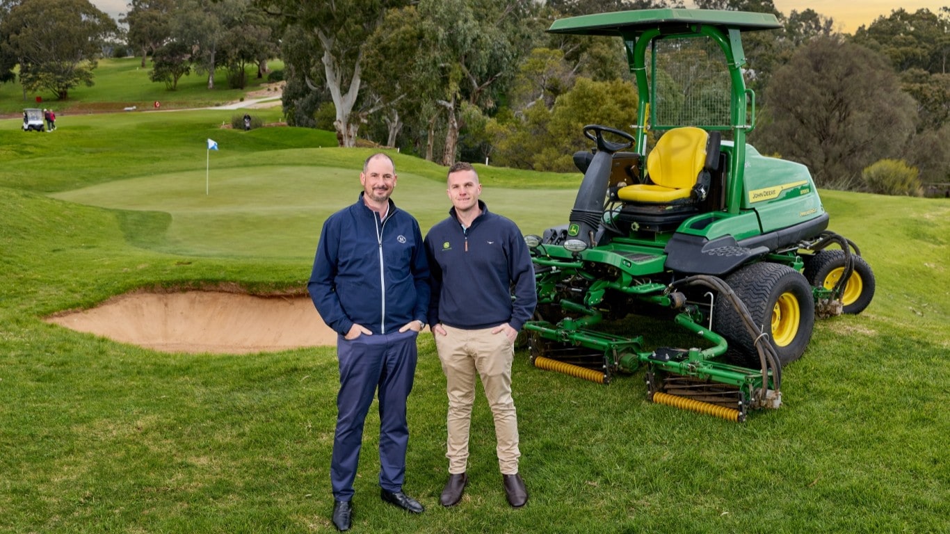 Two men with John Deere mower in front of golf green.