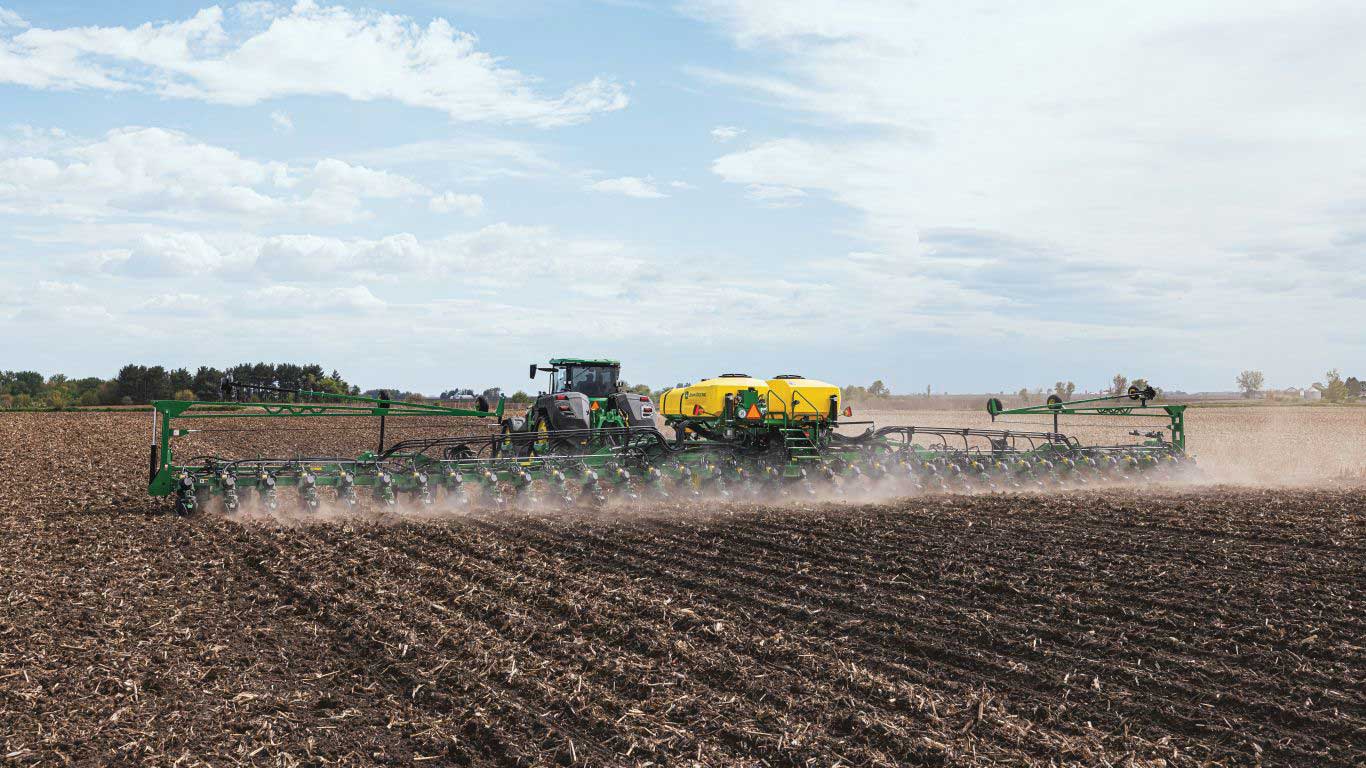 Green planting machine being pulled by tractor through soil with blue sky and clouds