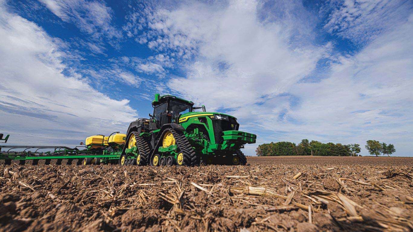 Ground view of large green tractor pulling planting machine with brown ground, blue sky with clouds and trees in the background