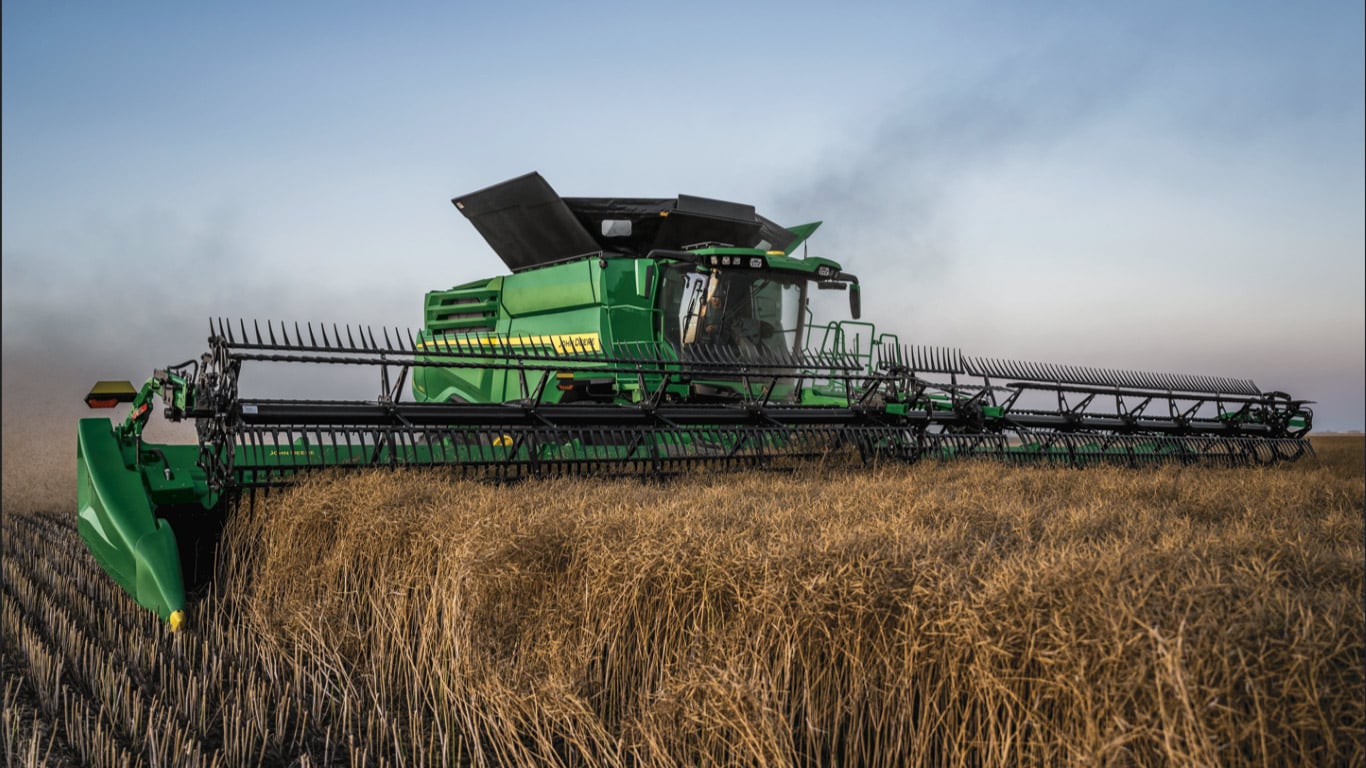 Large green machine with black spikes at the front harvesting crop with moody sky.
