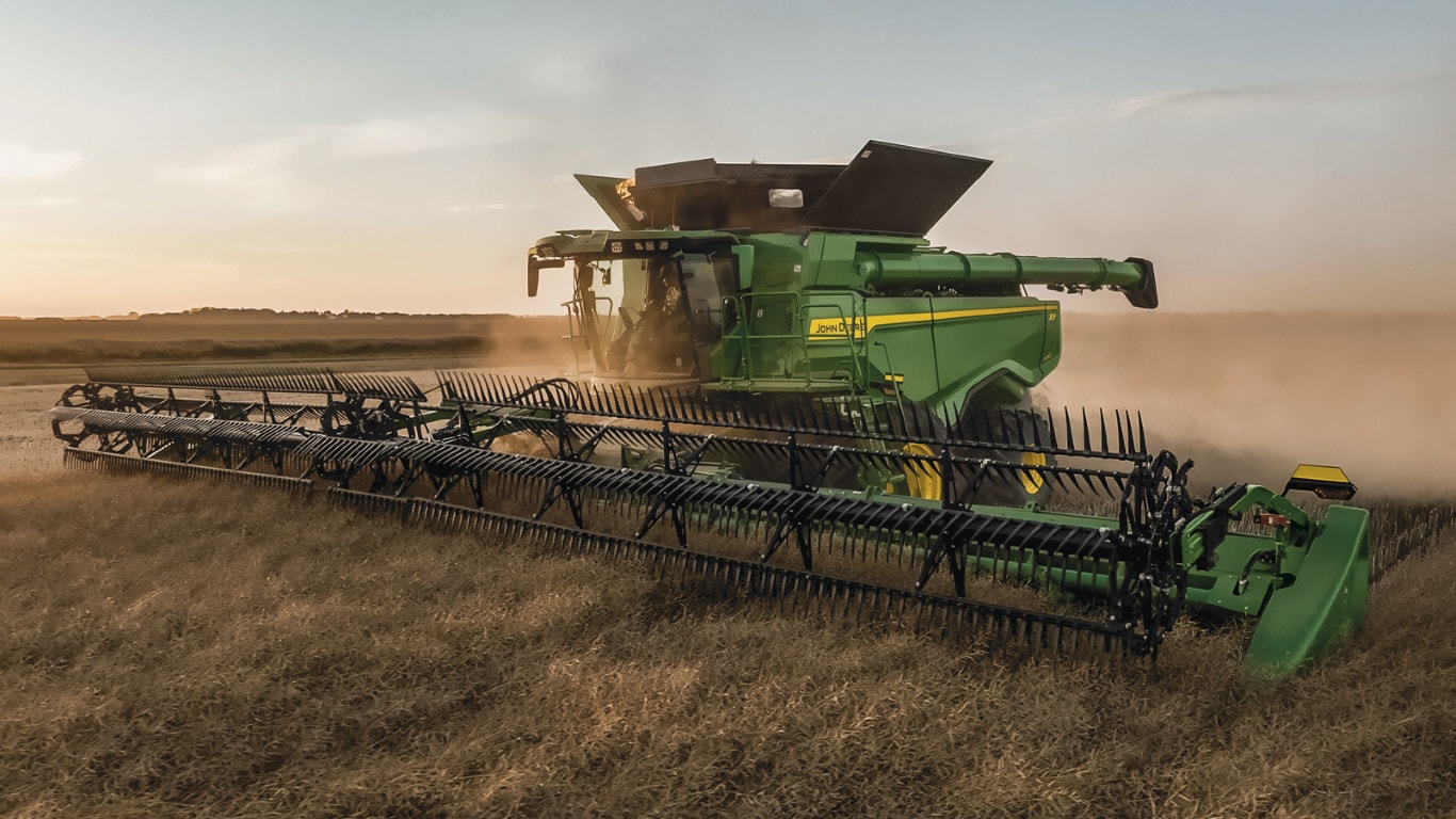 Large green machine with black spikes at the front harvesting crop during dusk