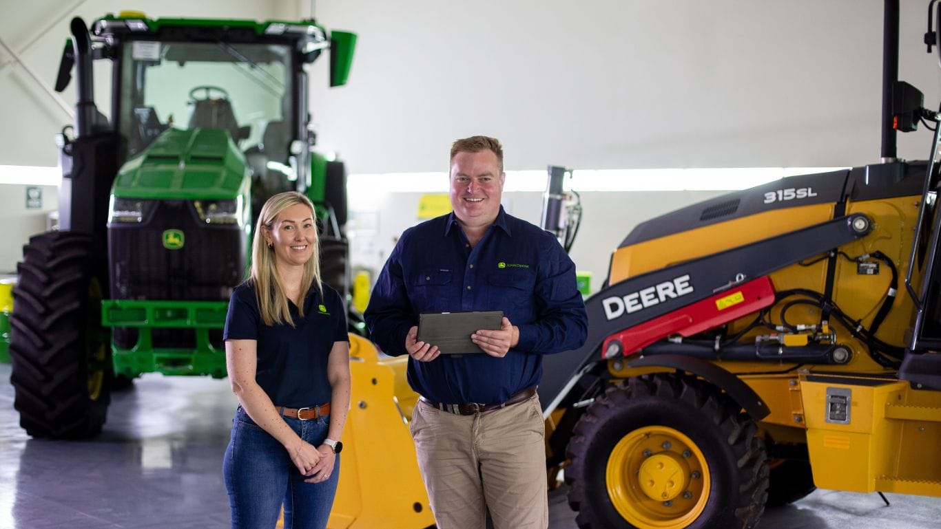 Two John Deere executives in a machinery workshop reviewing information on a tablet beside John Deere agricultural and construction equipment