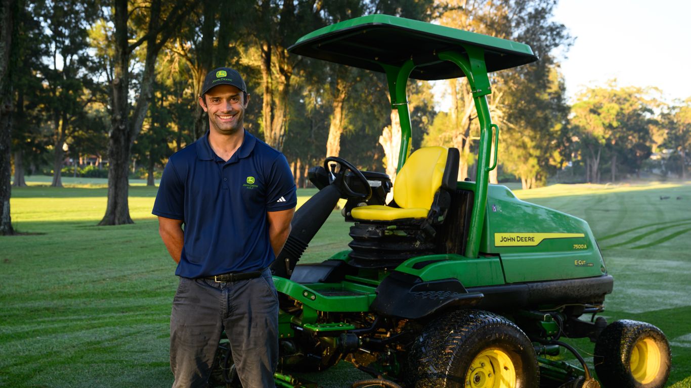 A man wearing a blue polo shirt and cap stands next to John Deere Golf equipment on a fairway.