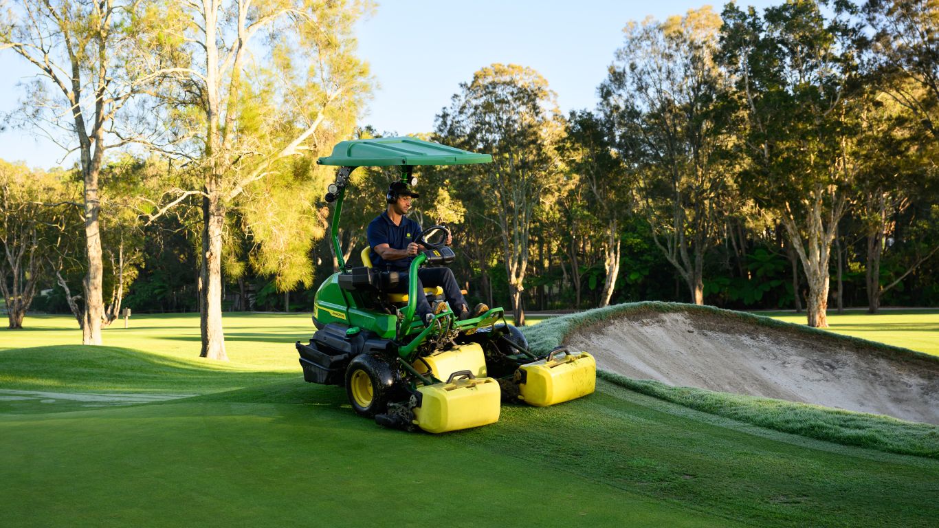 A man wearing a blue cap is driving a John Deere fairway mower on a golf course next to a bunker.