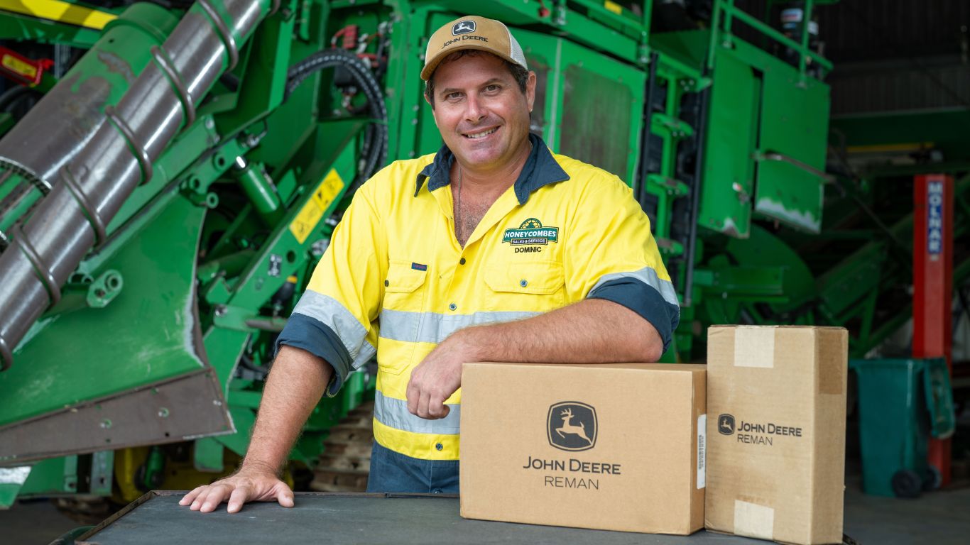 Dom Succio smiles in a workshop, leaning on Reman boxes in front of a John&nbsp;Deere harvester.