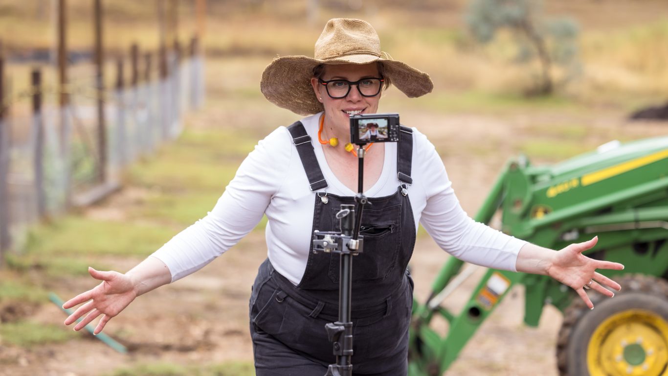 An image of a lady in a paddock in front of a camera and tripod and wearing a white top, overalls and a wide brimmed hat. Her arms are stretched out and she has a tractor behind her and a fence.
