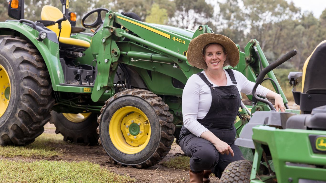 An image of a lady wearing a white top, overalls and a wide brimmed hat kneeling next to John&nbsp;Deere equipment.