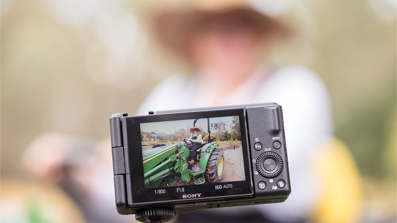 The background is a blurred picture of the woman on the tractor, with the foreground showing a close up of the back of a camera that shows the same image, although in focus, on the review screen.