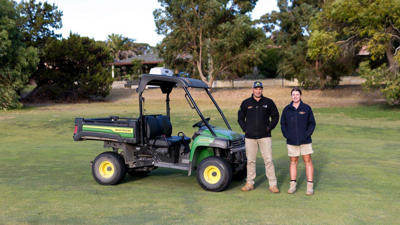 Two people standing beside a green utility vehicle on a golf course.