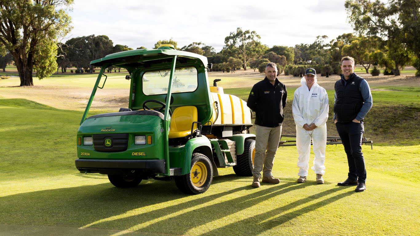 Three people standing beside a green utility vehicle with a yellow spray tank on a golf course.