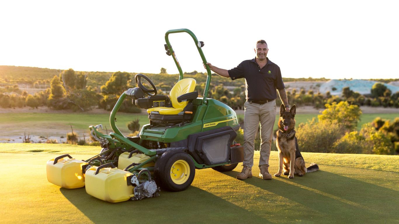 A person standing beside a mower with a German Shepherd on a golf course at sunset.