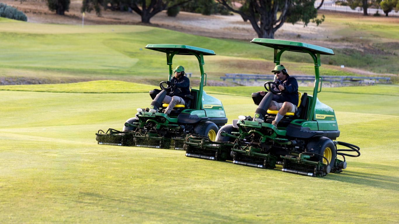 Two people driving green fairway mowers side by side on a golf course.