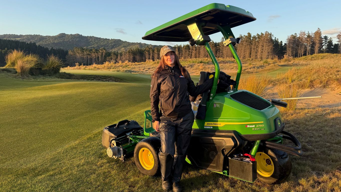 A lady with long brown hair wearing waterproofs standing next to a John Deere golf mower on a golf course.