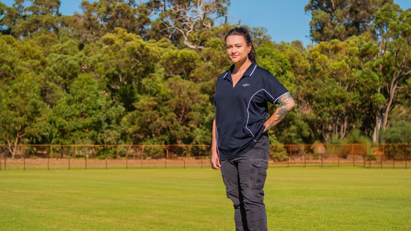 A lady wearing a blue polo and work trousers standing on a sports field with trees in the background.
