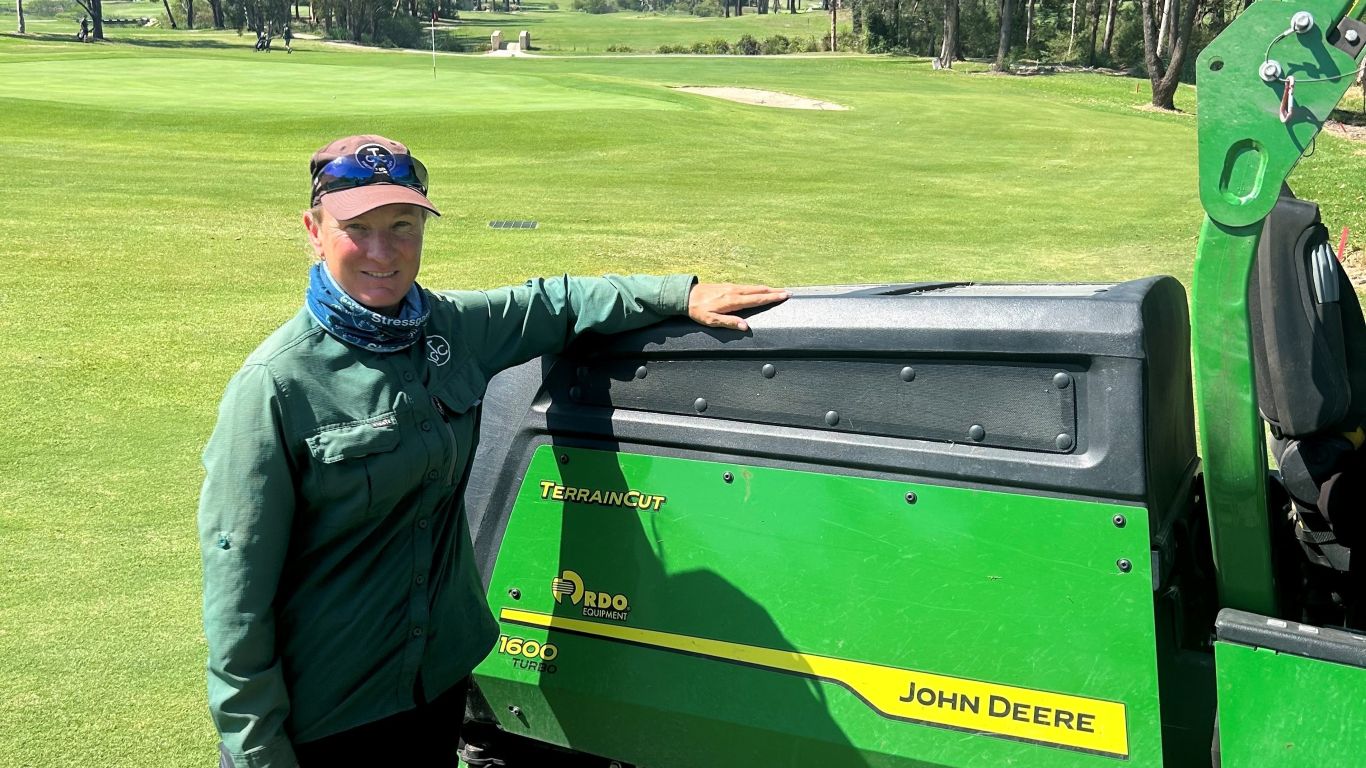 A lady wearing a green jacket and brown cap standing next to a John Deere terrain cut mower on a golf course.
