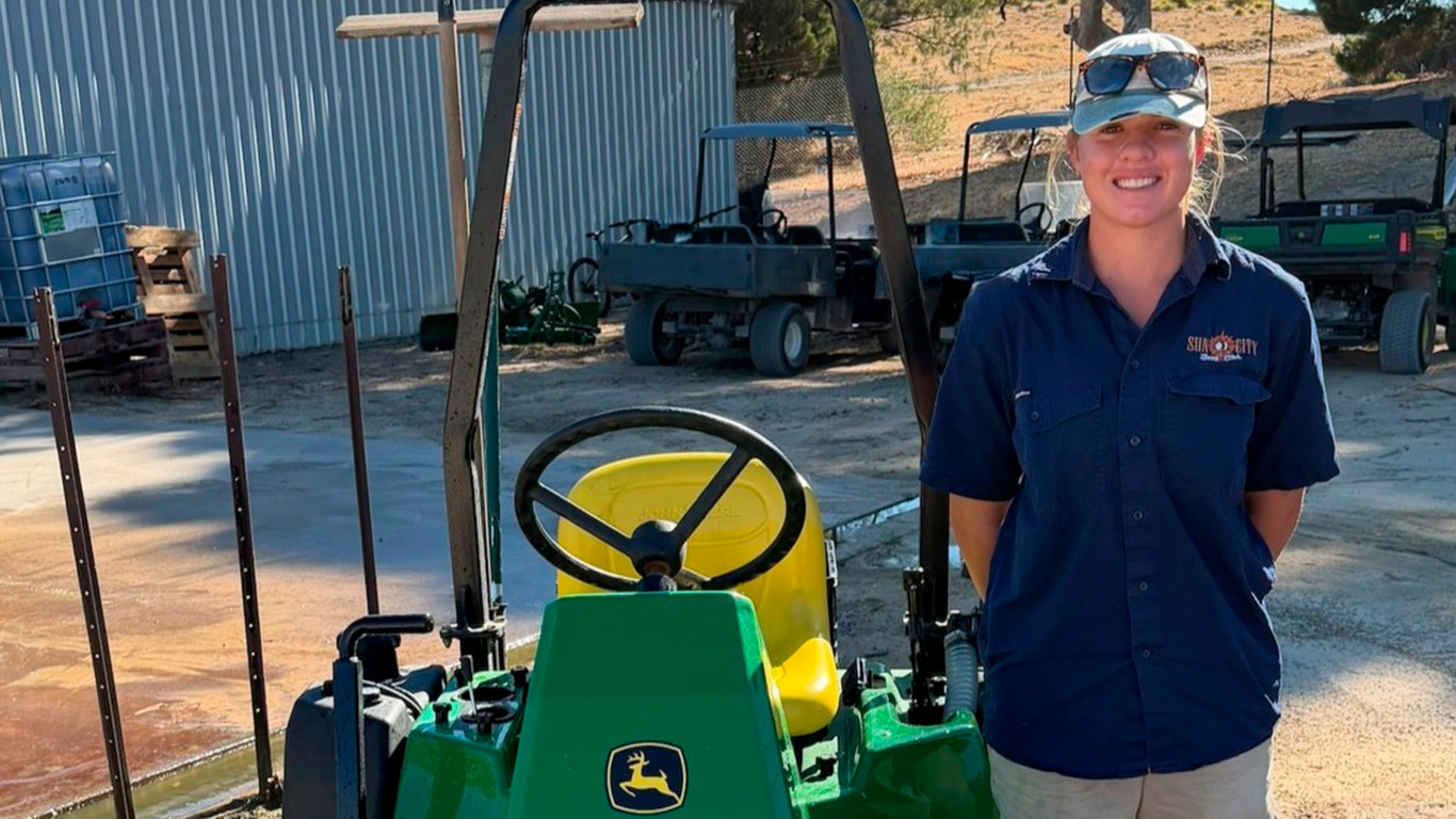 A lady wearing khaki shorts and work boots, with a blue polo shirt and cap stands next to a bunker rake.