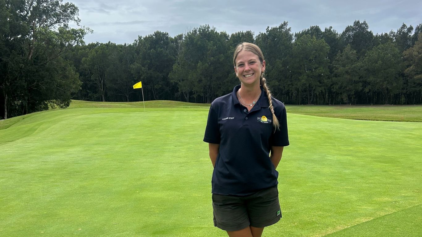 A lady with a long blonde plait standing on a golf course fairway wearing a navy blue polo shirt and black shorts and work boots.