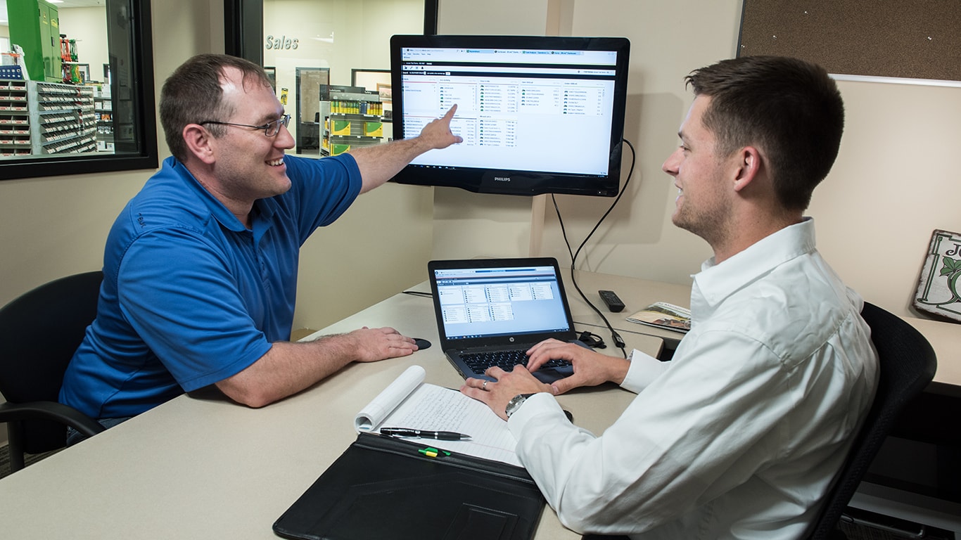 a man at a desk pointing to a monitor while talking to another man