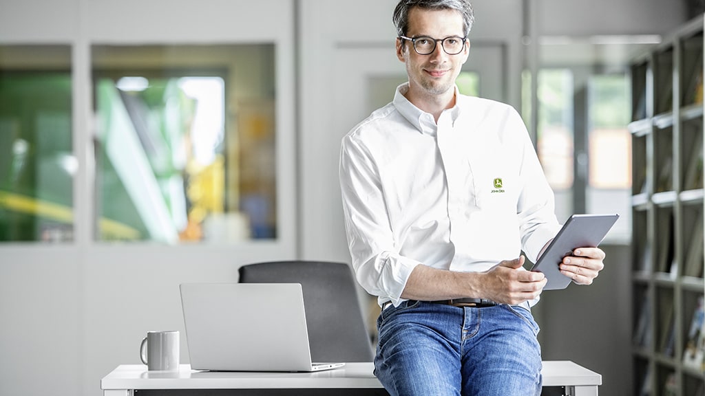 a man in a John Deere shirt leaning on the front of a desk in an office and holding a tablet
