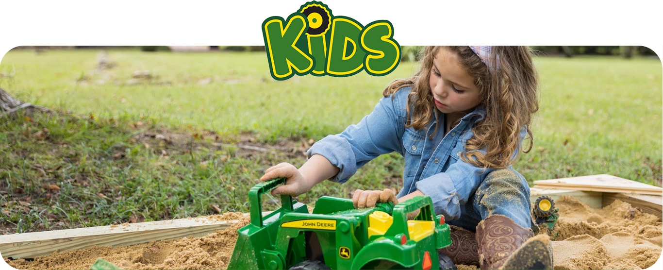 Young girl playing with John&nbsp;Deere toy tractor in sandbox around grass