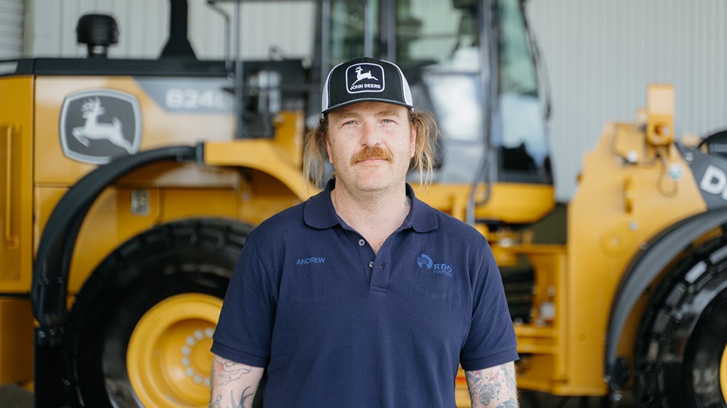 Andrew Moat wearing a black and white cap, standing in front of a large 824 Wheel Loader, smiling to camera.
