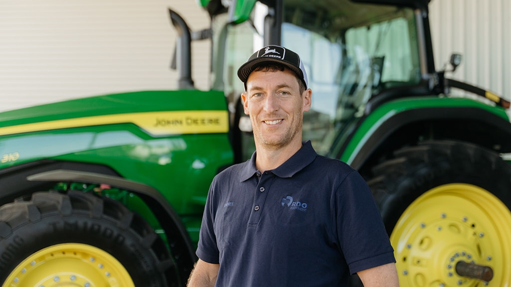 Daniel Barnes wearing a black and white cap, standing in front of agriculture equipment, smiling to camera.