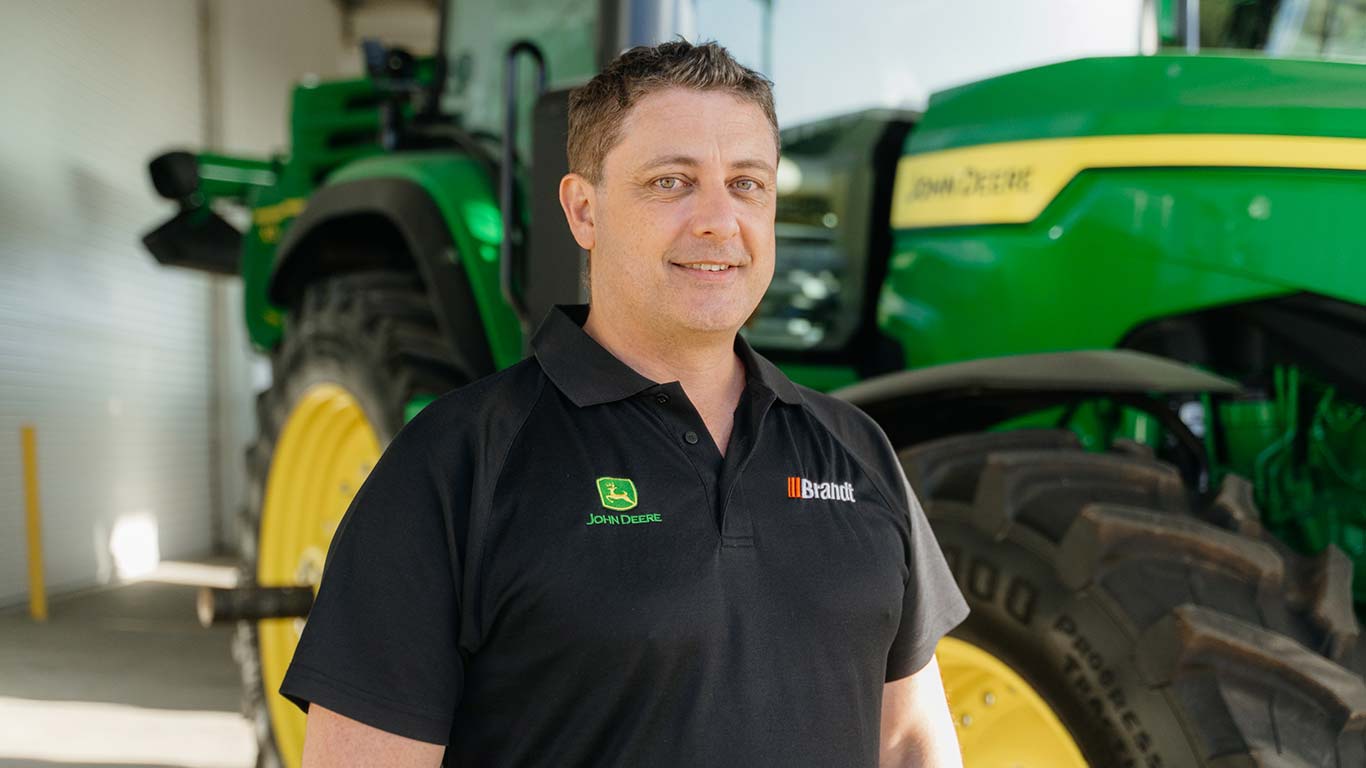 Daniel Stoneley standing in front of green agriculture equipment, smiling at the camera.