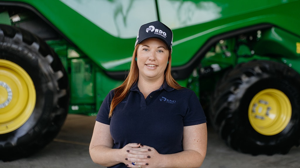 Tracy Hogan standing in front of large agriculture equipment, smiling to camera.