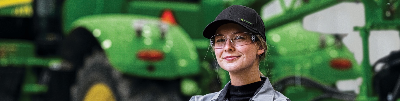 Grinning woman with safety glasses standing in front of an out of focus tractor