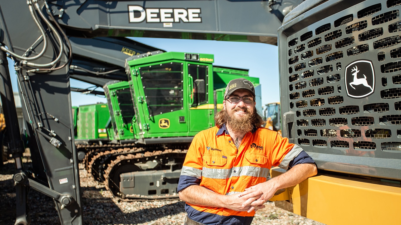 Brendan smiling with hands clasped while leaning Deere forestry equipment