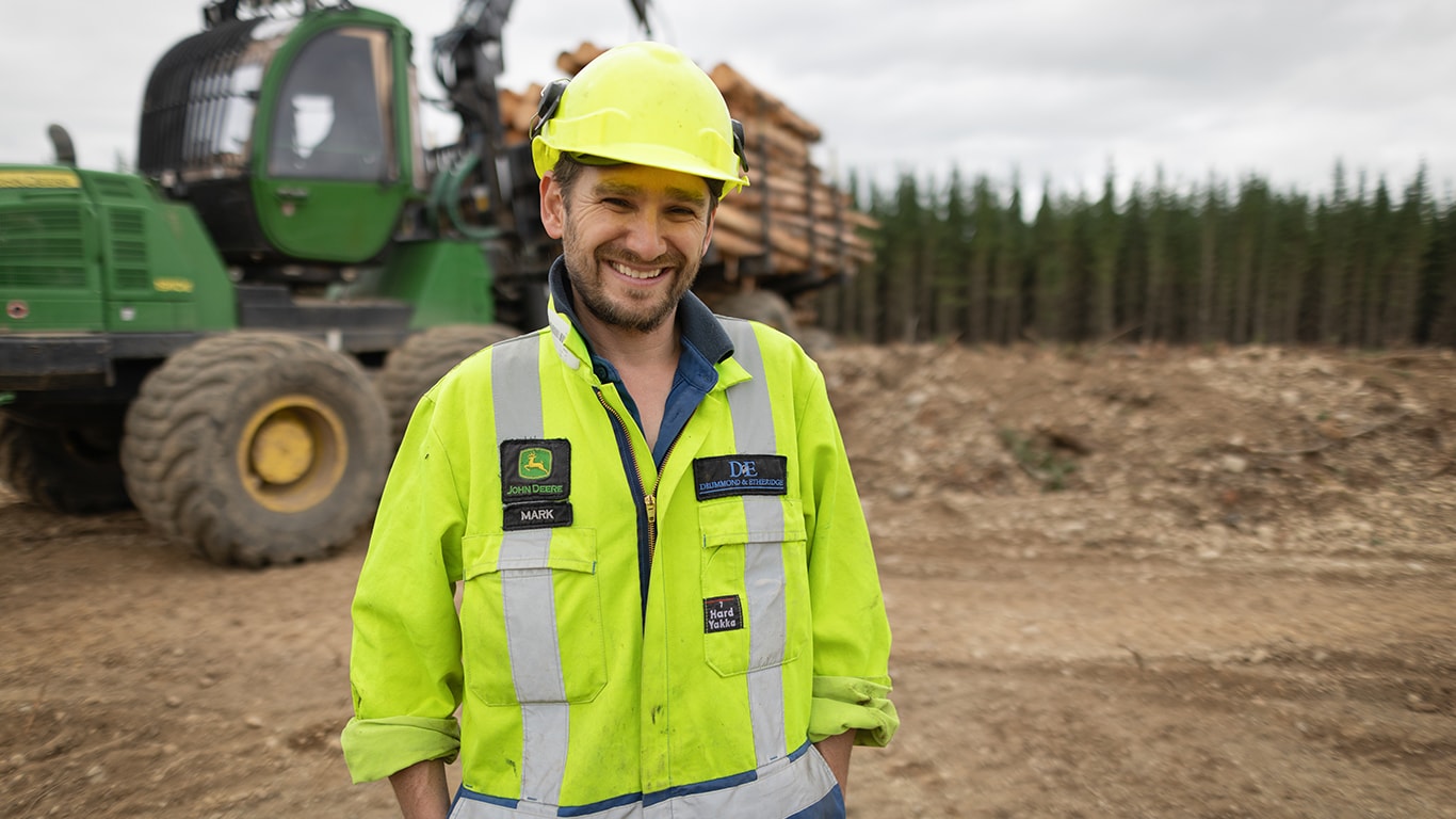 Mark smiling with his hands in his pockets and forestry equipment behind him