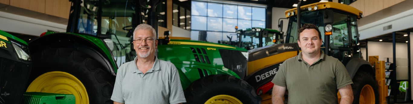 two smiling people standing in front of a John Deere 6175M tractor and a 315SL Backhoe in a showroom