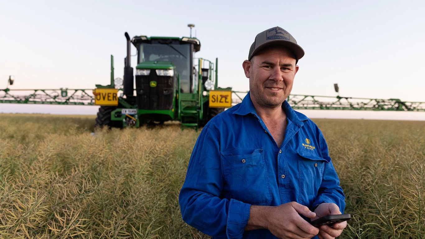 An Australian customer is in his field smiling direct to camera. In the background is his self-propelled sprayer with the booms fully extended.