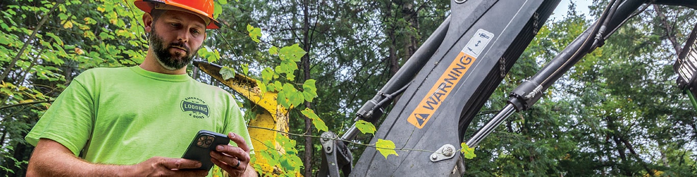 a forestry worker with orange hardhat and flourescent green tshirt on interacting with a mobile phone in the forest