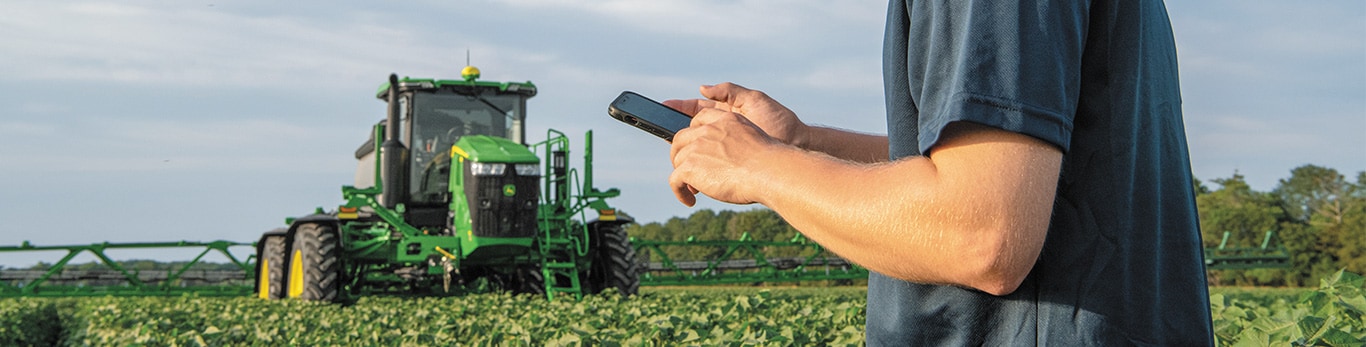 closeup of farmer's forearms interacting with a mobile phone with a John Deere tractor in a field of leafy crops