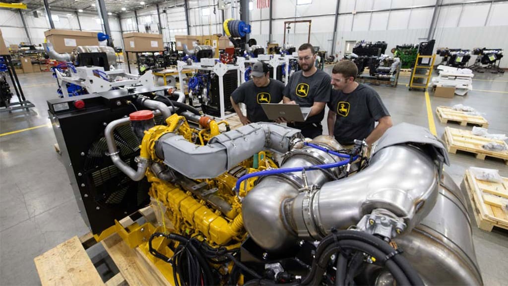 Large yellow engine on a pallet with three people reviewing data on a laptop in a factory.