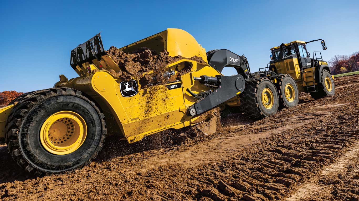 Yellow earthmoving scraper and tractor dumping soil on a dirt construction site under a clear blue sky.