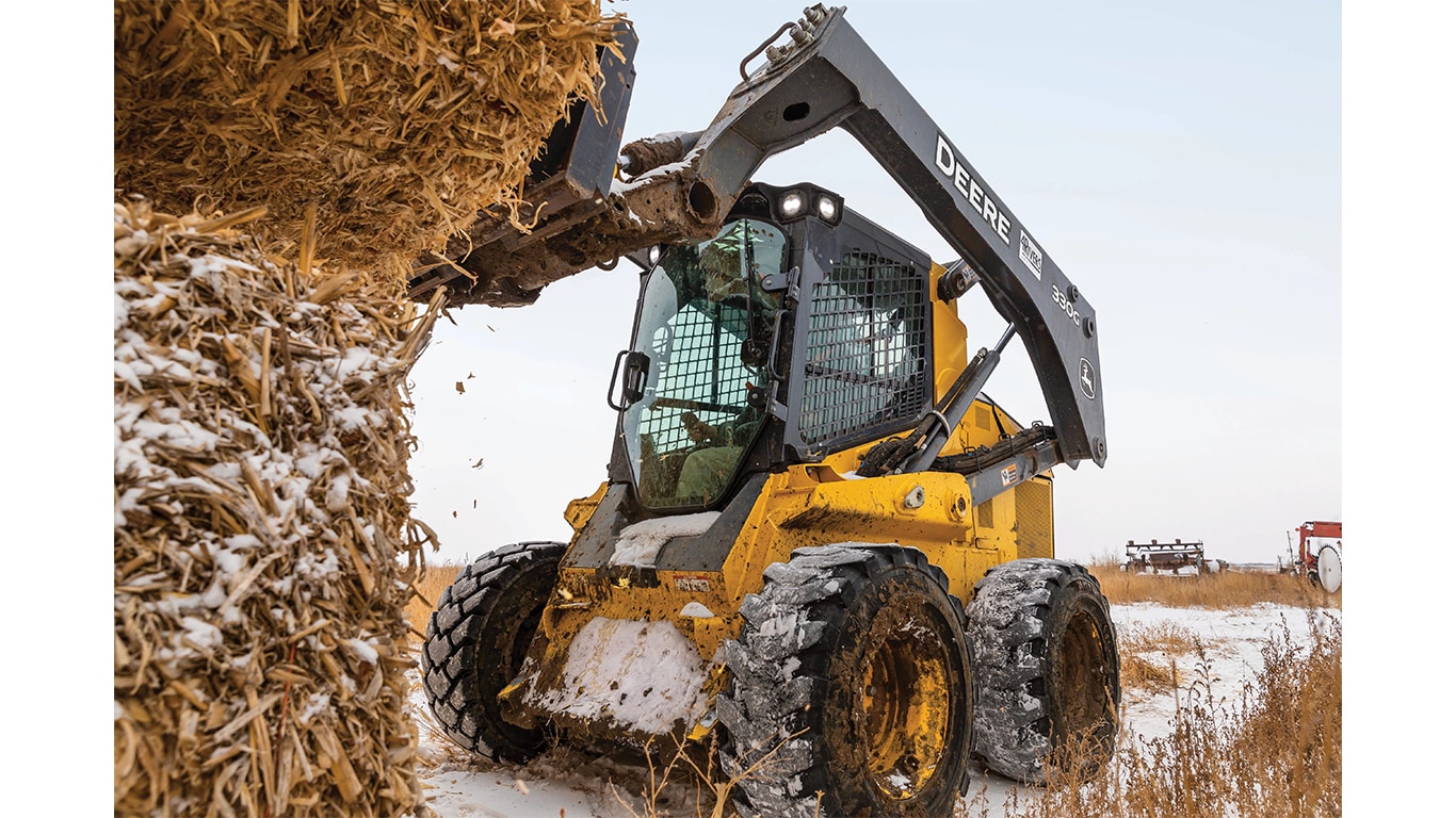 A 330G Skid Steer with bale spears attachment lifting a bale of snowy hay in a snowy field.