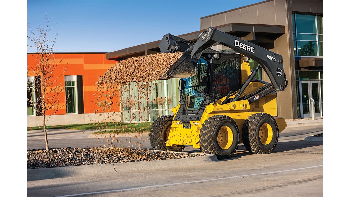 A 330G Skid Steer dumping rocks in a parking lot landscape area with buildings in the background.