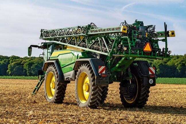 John Deere 500R self‑propelled sprayer parked on a harvested field, rear view with spray boom folded for transport.